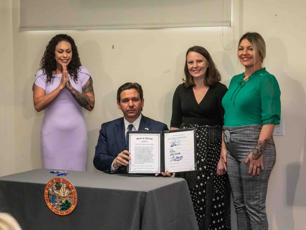 A seated Gov. Ron DeSantis, which three women identified as victims of Jeffrey Epstein, Hayley Robson, Jena-Lisa Jones, and Courtney Wild, standing alongside him, holds up a bill he just signed that will allow for the release of grand jury testimony from a 2006 case investigating the pedophile financier.