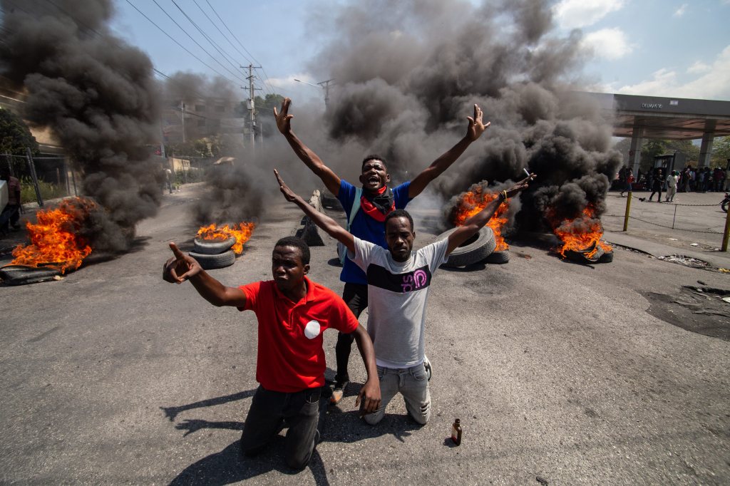 Three Haitian men hold their arms up in defiance on a street littered with burning tires in Port-au-Prince.
