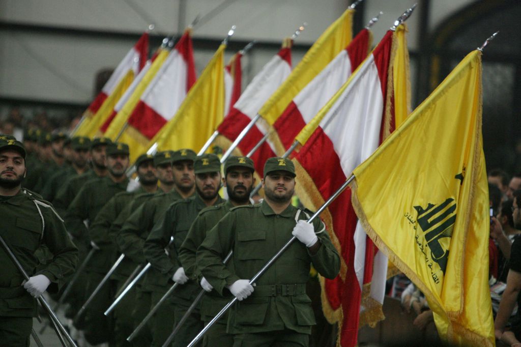 A rank of Hezbollah fighters in green uniform and white gloves holding across their chests flagpoles. Each flagpole carries alternately the Hezbollah yellow flag which is green Arabic script on yellow, and the Lebanese national flag. 