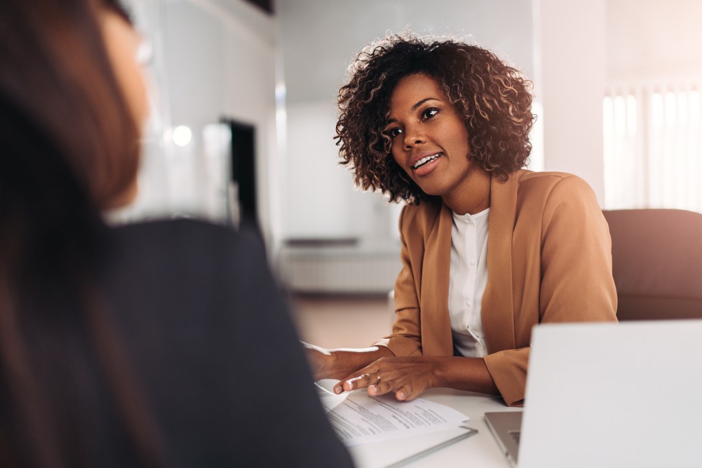 Young woman doing job interview in office talking with client. Laptop and papers on table.