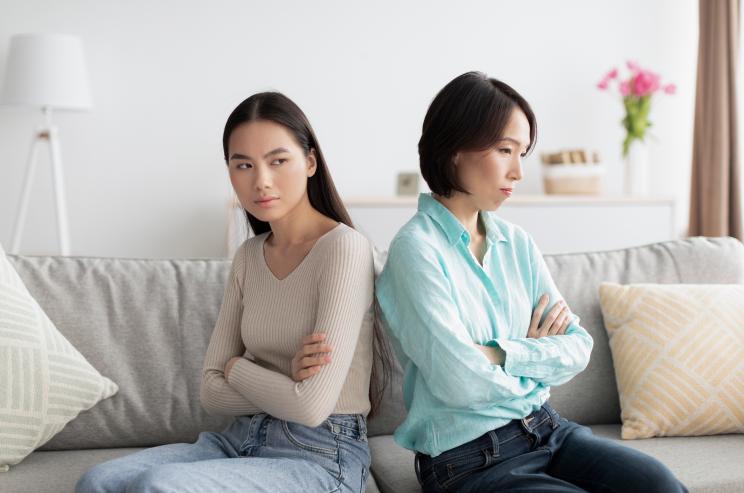 Offended Asian mother and daughter sit back-to-back on a couch ignoring each other after an argument in their home - iStock-1468483264.