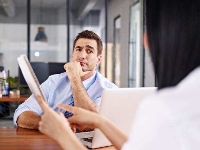 a skeptical caucasian male interviewer listening to an asian female interviewee; conversation happening over a laptop