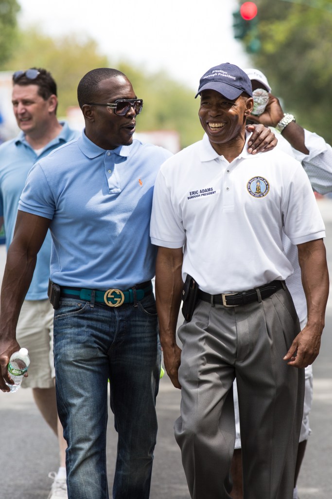 Lamor Whitehead and Eric Adams walking. Whitehead has his arm around Adams and is wearing a blue polo, jeans and a Gucci belt. Adams is wearing a white polo with his name and gray pleated slacks. Adams is smiling broadly and Whitehead appears to be speaking to him