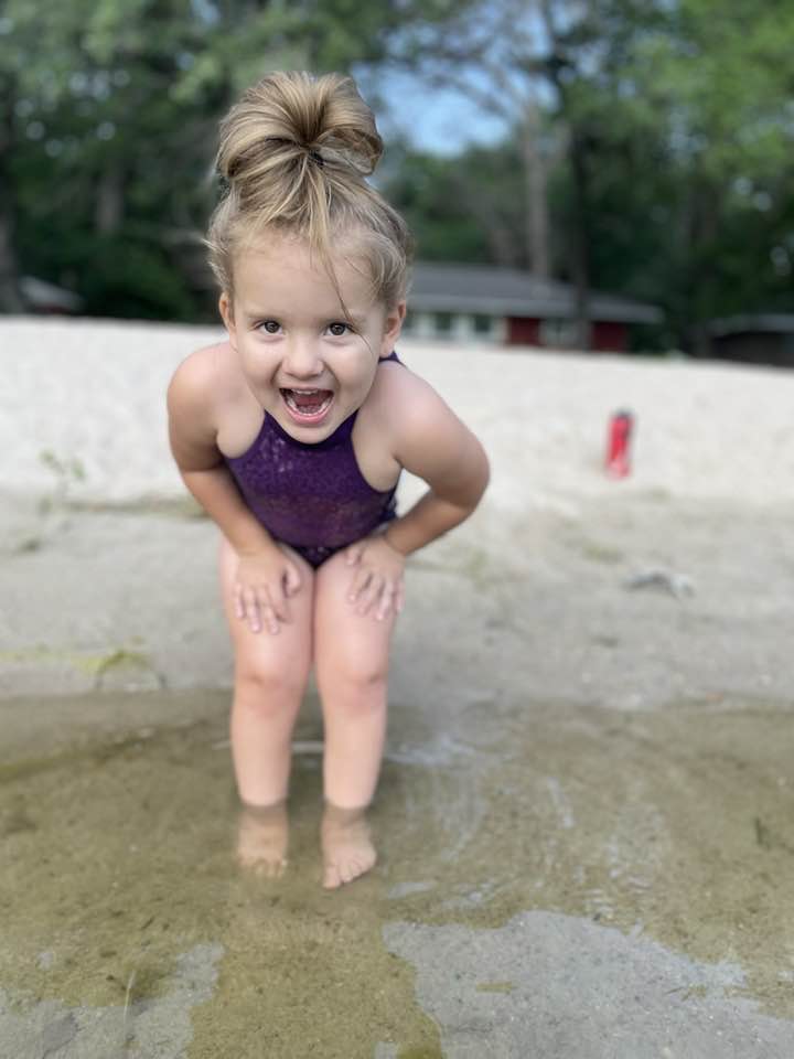 A little girl in a purple bathing suit crouches with her feet in a body of water.