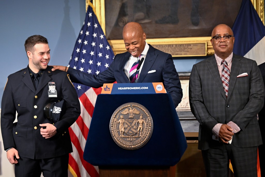 Mayor Eric Adams smiles and puts his arm around a young NYPD officer in full uniform. To the right in a checked suit is Philip Banks, deputy mayor for public safety.