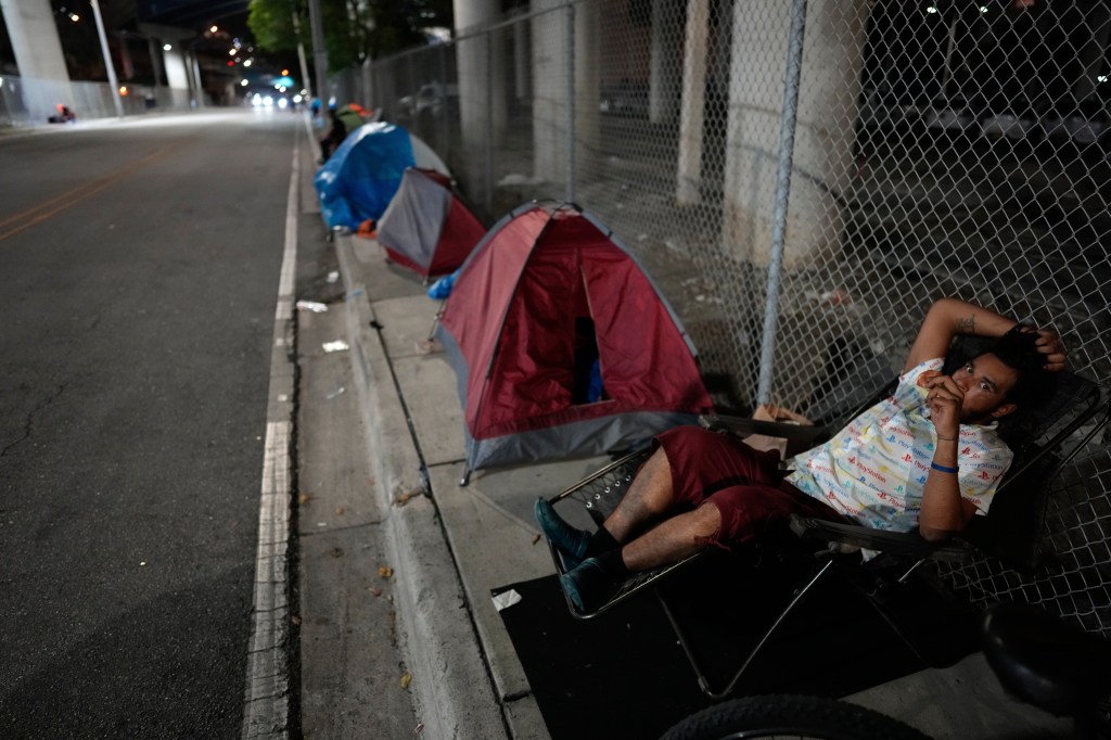 A person rests in a lounge chair on a sidewalk alongside tents as a team from the Miami