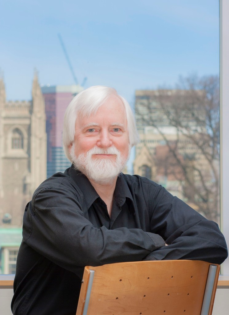 Joe Carens sits backward in a chair with college buildings as a backdrop. 