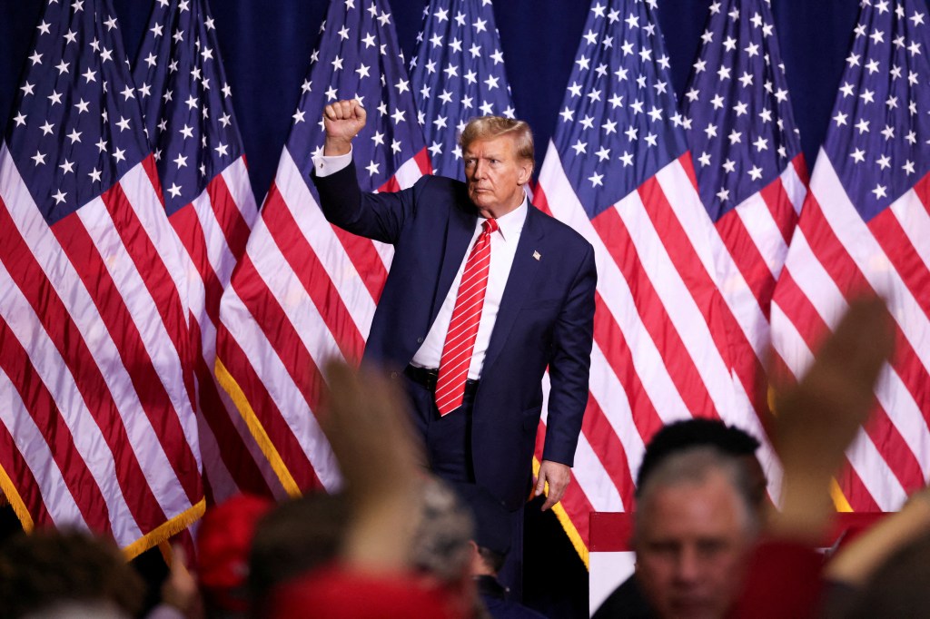 Former U.S. President Donald Trump reacting during a campaign rally, standing in front of a crowd of flags at the Forum River Center in Rome, Georgia.