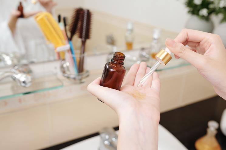 Close-up of a woman applying serum of facial oil from a dropper in front of a bathroom mirror