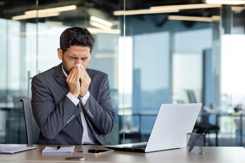 A man sneezes into a tissue while using a laptop at work.