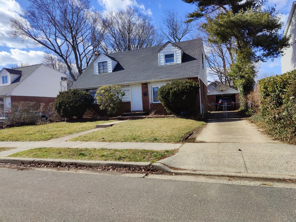 The two story home with white front door, boarded up front window, front lawn and driveway.