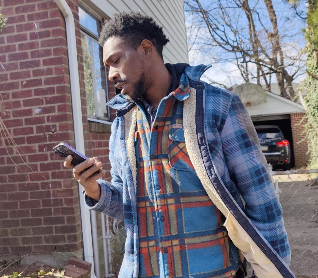 denton gayle in blue plaid coat over plaid shirt looking at a cell phone in his hand in the driveway of 39 brussel dr