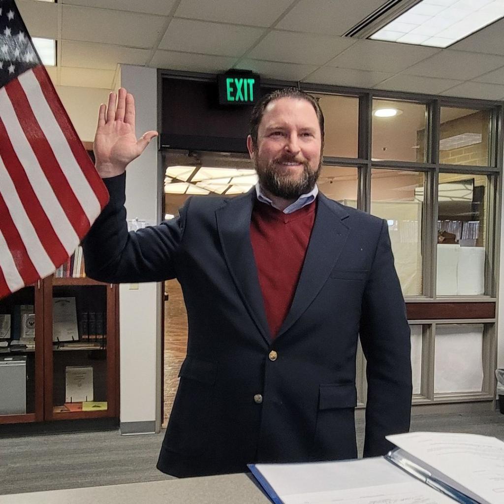 Utah State Representative Brett Garner raising his right hand, while standing in front of an American flag.