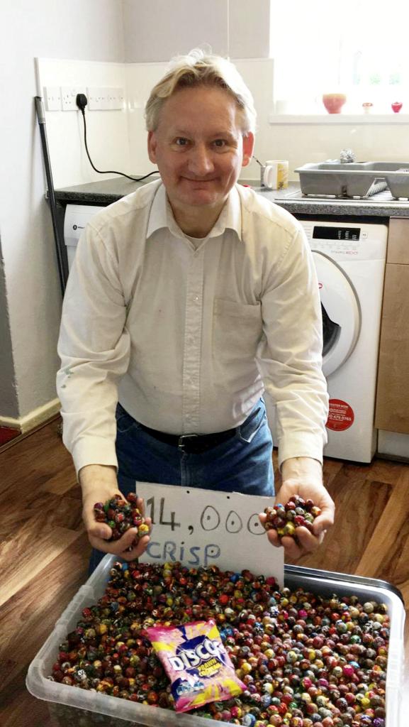 Man holding sign with a price tag in front of collection of empty crisp packets.