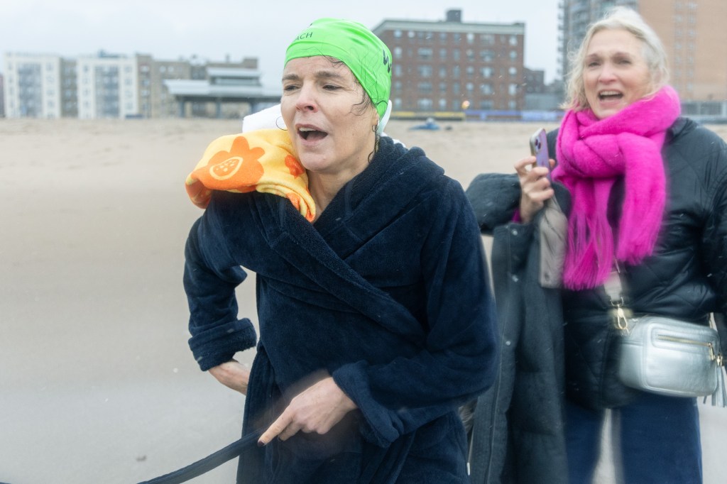 Wall Street Journal Editor-in-Chief Emma Tucker, wearing a bright green bathing cap, wraps a blue robe around her after swimming in the frigid water off Brighton Beach to call attention to detained reporter Evan Gershkovich's case. Behind her, a supporter wearing a bright pink scarf smiles.