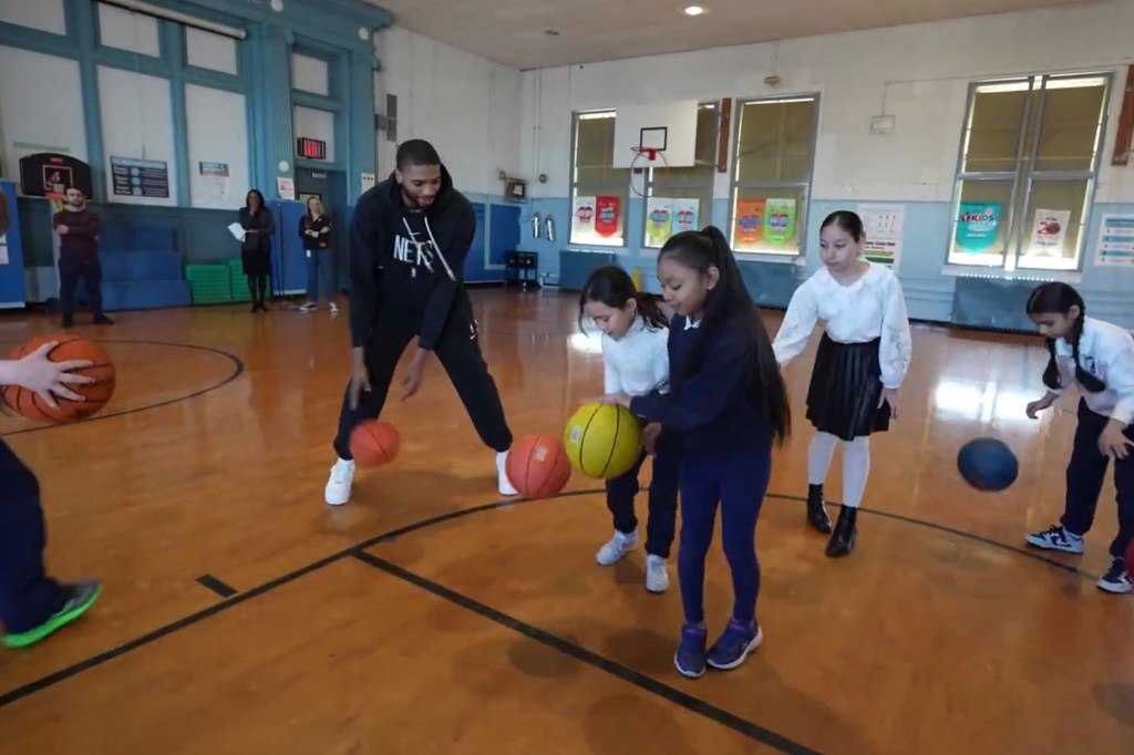 Mikal Bridges dribbles a basketball in a school gym with young students. 