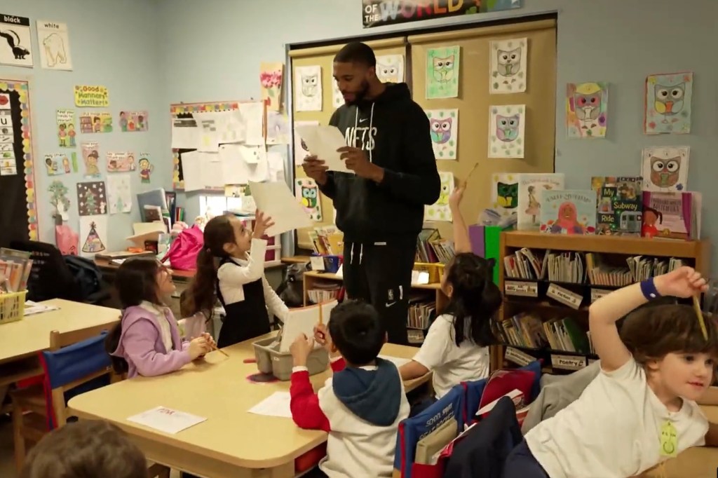 Mikal Bridges looks at a  paper as he stands at a table with four students seated in a classroom. 