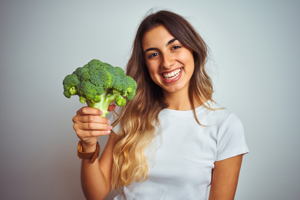 Young beautiful woman eating broccoli over grey isolated background with a happy face standing and smiling with a confident smile showing teeth