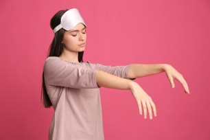 Young woman sleepwalking in pajamas and sleep mask against a pink background