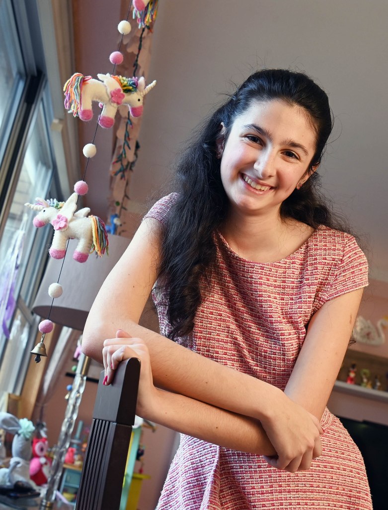 14-year-old Kristina Raevsky smiling at the camera in her Forest Hills apartment, leaning on chair, wearing pink dress.