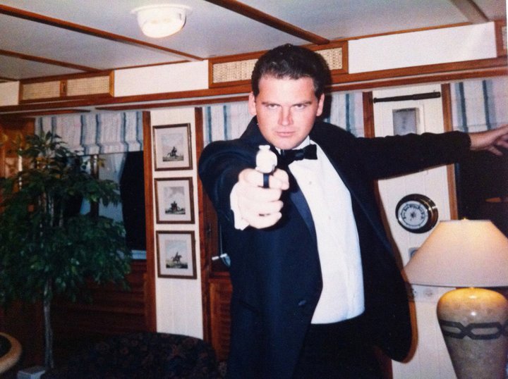 Andrew Getty in a tuxedo aiming what appears to be a toy gun at the camera, in front of wooden decor and paintings. 