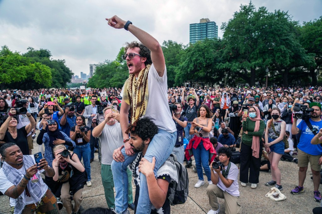 Ammer Qaddumi participating in a chant outside the Main Building on UT-Austin campus during a protest, surrounded by a crowd of professors, students, and supporters