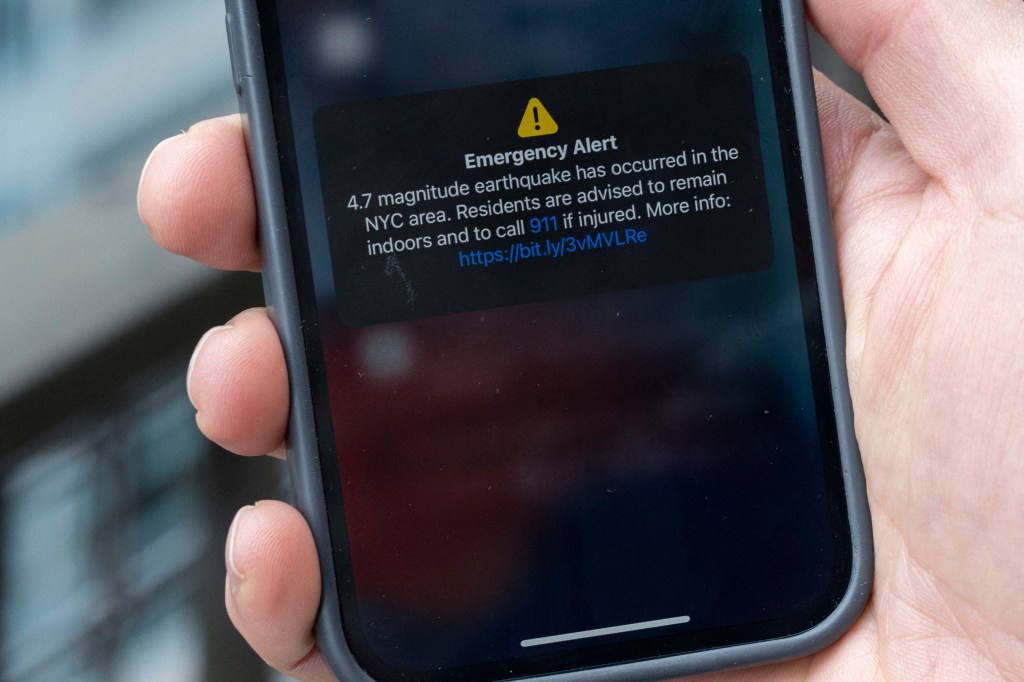 A smartphone held on a person's hand displaying an Emergency Alert message about the earthquake in New York City.
