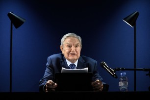 George Soros, in a suit, addressing the assembly with a microphone and water bottle at the World Economic Forum annual meeting in Davos, 2022