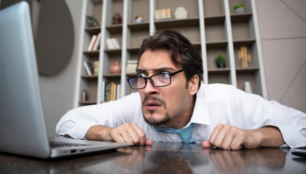 Shocked businessman hiding behind desktop and looking at laptop in the office. Business crisis and bankruptcy concept