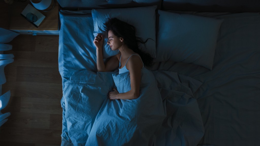 Top View of Beautiful Young Woman Sleeping Cozily on a Bed in His Bedroom at Night. Blue Nightly Colors with Cold Weak Lamppost Light Shining Through the Window.