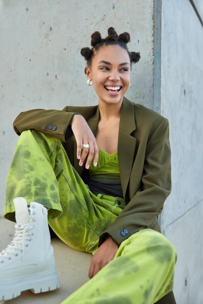 Positive carefree teenage girl with bun hairstyle looks happily away wears stylish green costume and white boots blue eyeliner feels good poses against grey concrete wall. Urban lifestyle concept