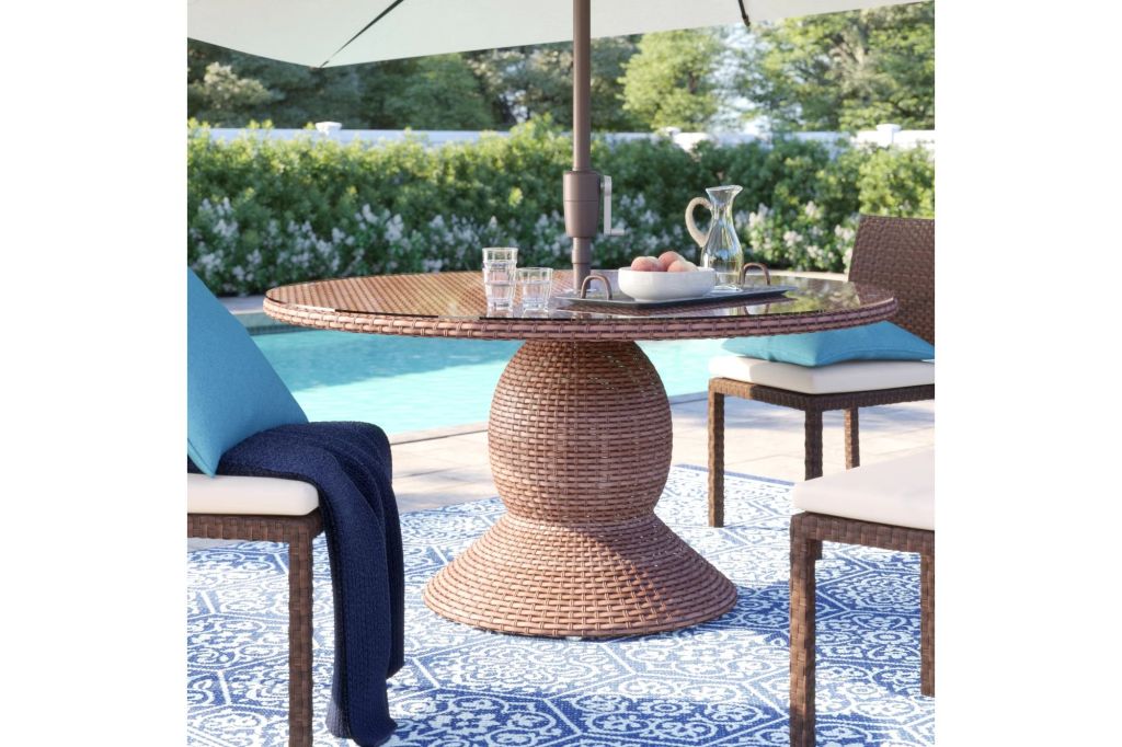 A round wicker coffee table with a glass top and an umbrella by a pool.