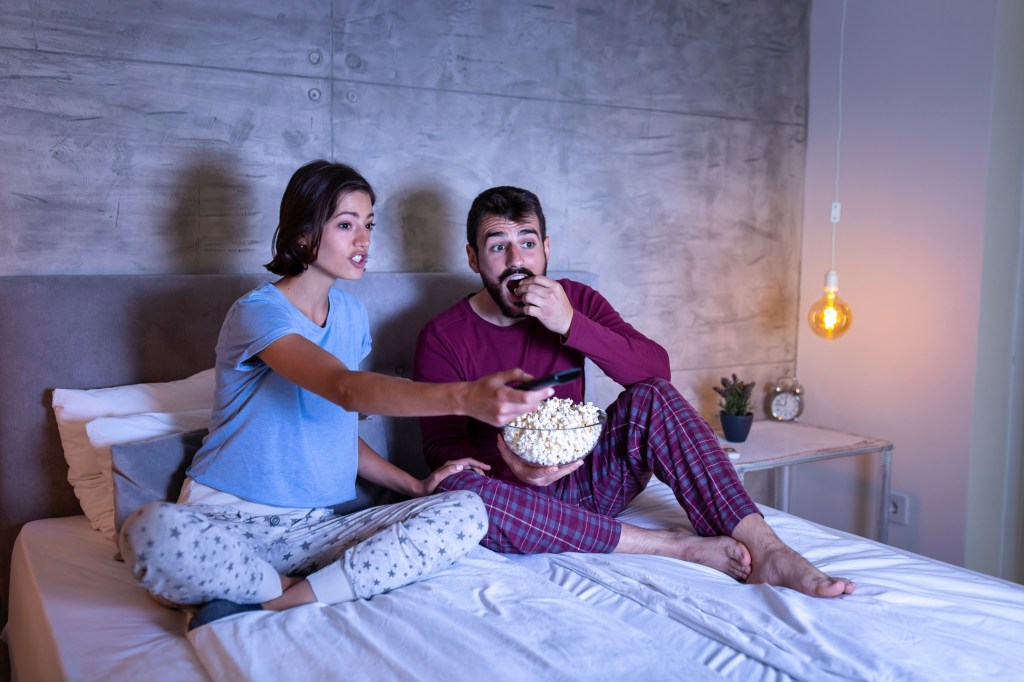 Beautiful young couple in love, sitting in bed side by side, eating popcorn.