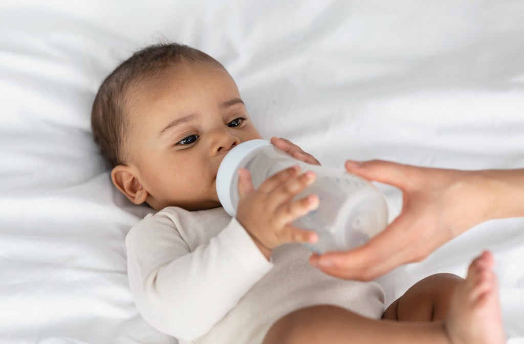 Woman helping a cute little African American baby drinking milk from a bottle while lying on a white blanket at home