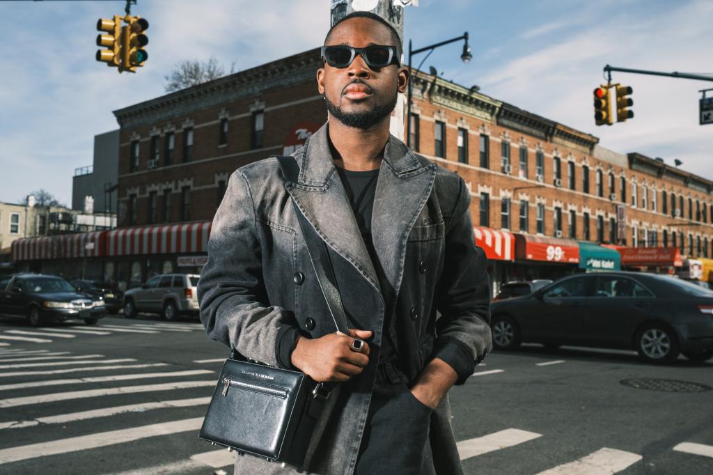 CJ Robinson in Brooklyn, NY standing at an intersection in a black trench, black glasses and black bag