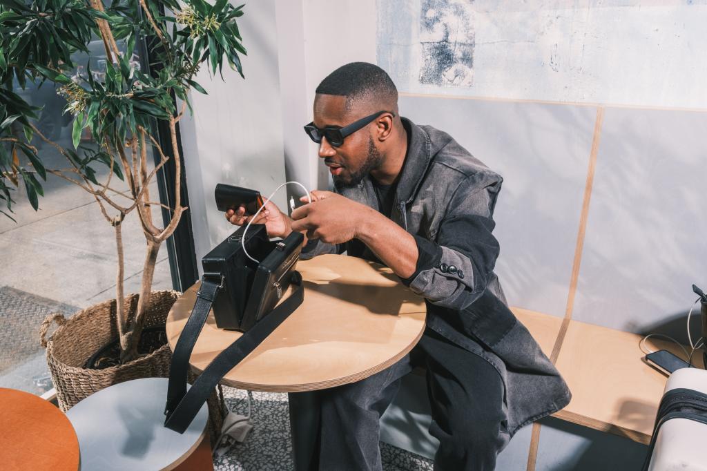 CJ Robinson, 28-year-old art director and illustrator, sitting at a table in Little Roy Coffe Co, Flatbush, Brooklyn, looking at his phone and carrying a Jordanis handbag by Brandon Blackwood