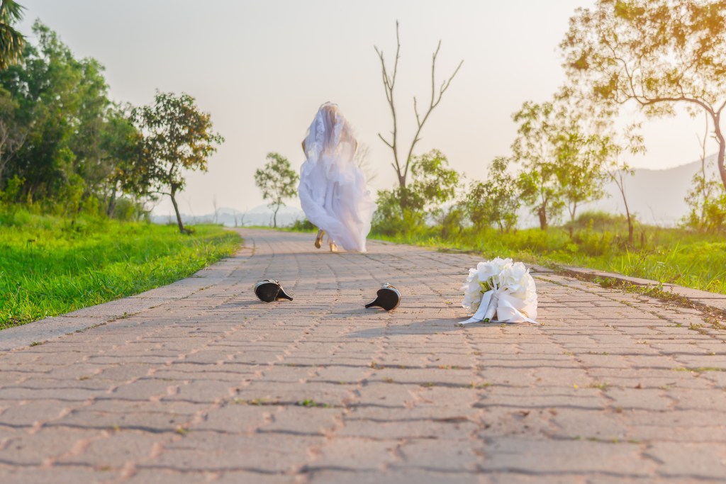Bride in white wedding dress running in nature, leaving behind a bouquet and shoes on a street