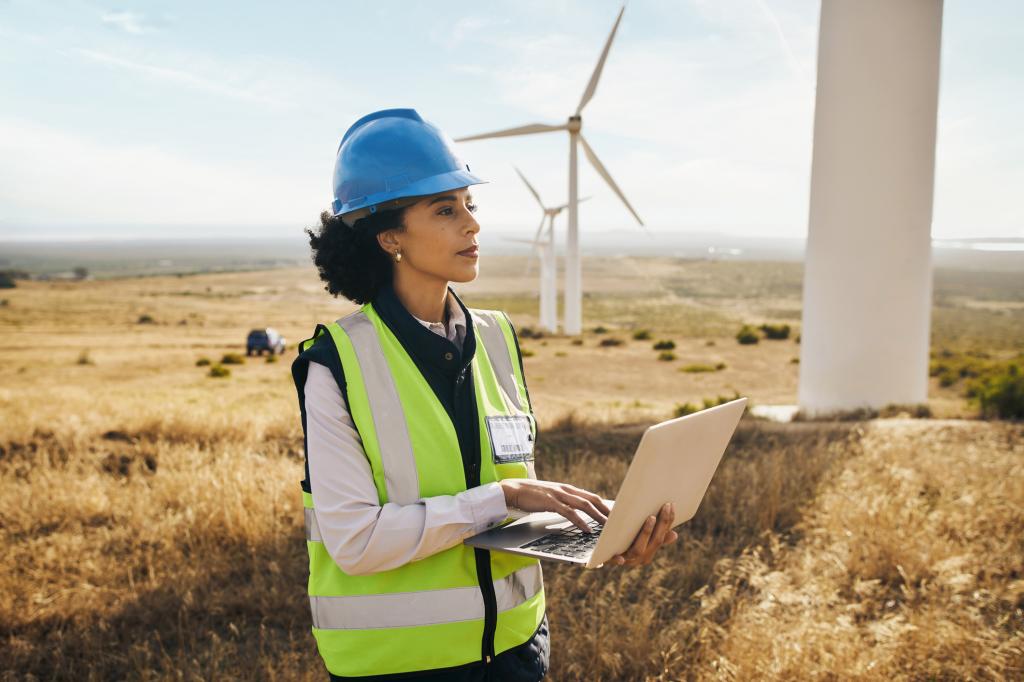 Black female engineer in safety gear typing on laptop, with wind turbines on African farm in background