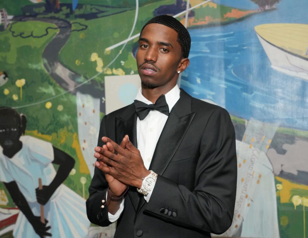  Christian Combs is wearing a tuxedo and  rubbing his hands together with a blank expression on his face