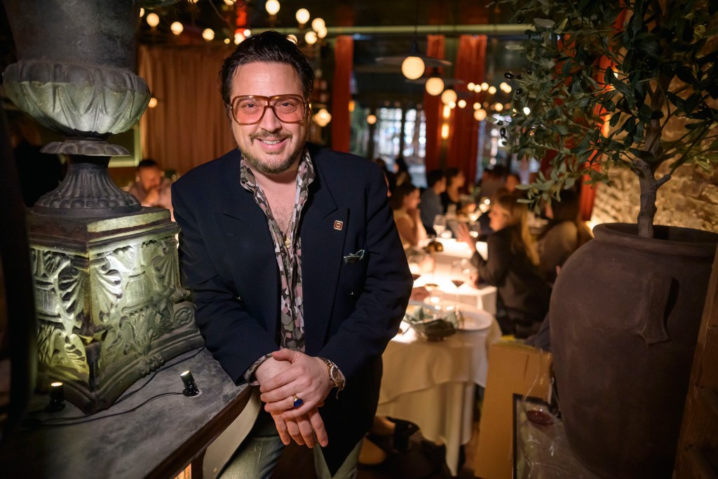 Max Tucci in tinted glasses, stands and smiles at Tucci, with out-of-focus diners behind him. 