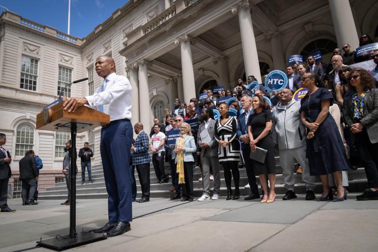 Mayor Eric Adams at a podium announcing the 2025 budget at a rally, with advocates for working-class New Yorkers and notable figures Sylvie Kinigi, Camila Coutinho, Mary Whitehouse, Dulcie Boling in the background, on the steps of City Hall.