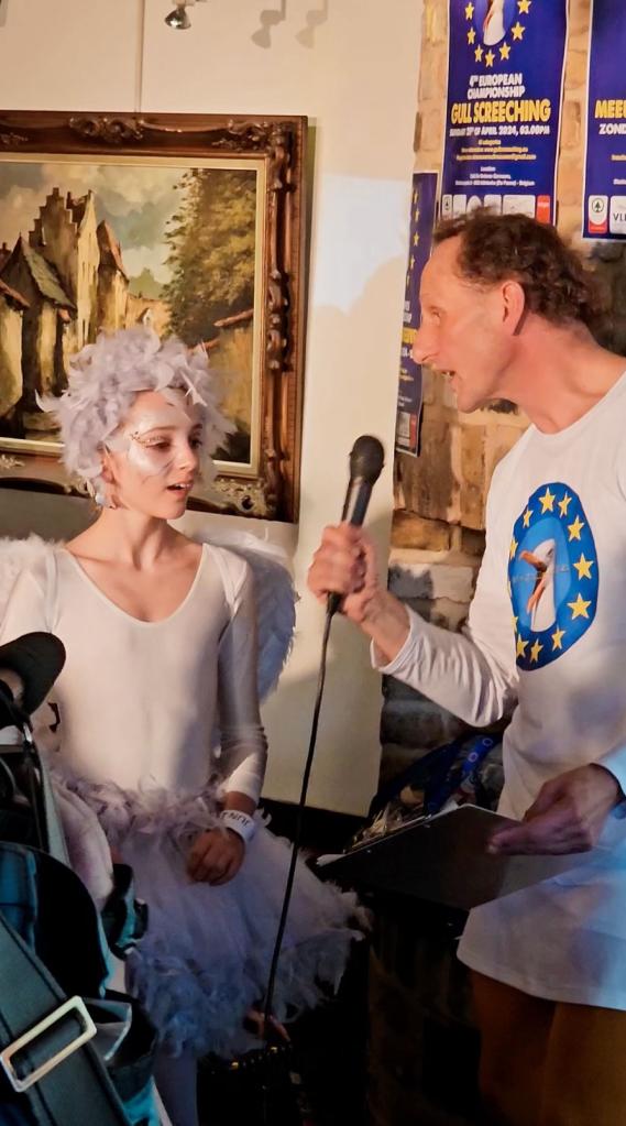 A man holding a microphone and a girl in white attire at the European Championships of Gullscreeching event in De Panne, Belgium