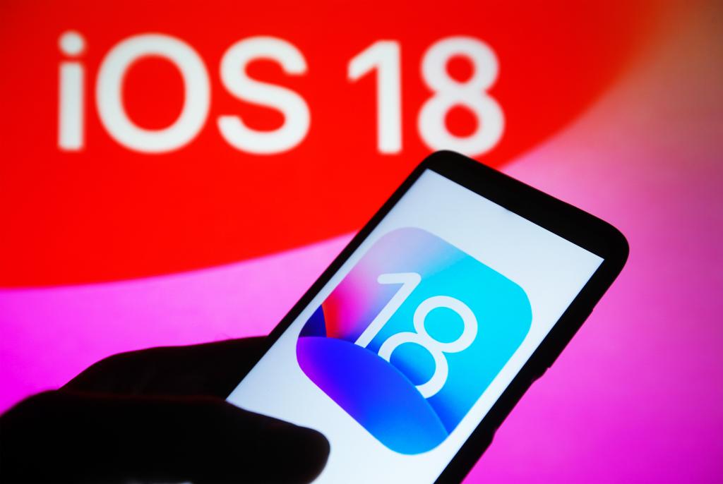 iOS 18 logo displayed on a smartphone with the logo also visible on a PC screen in the background