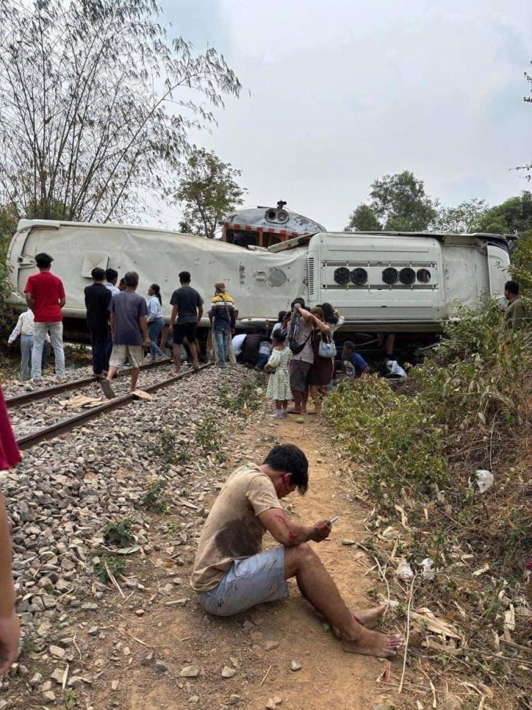 Train crashing into a white bus at a level crossing in Cambodia, with bystanders standing nearby