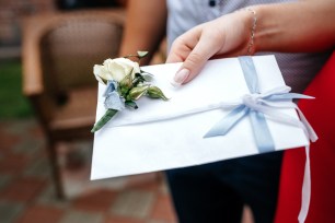 Woman's hand holding a white wedding envelope adorned with boutonniere, a gift from wedding guests