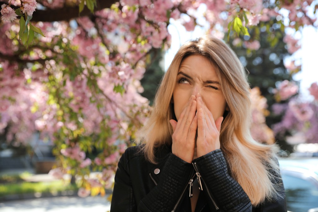 Woman covering her mouth with her hands due to seasonal pollen allergies near a blossoming tree outdoors