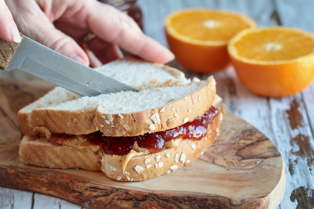 Woman's hand cutting a peanut butter and strawberry jelly sandwich on a rustic wooden cutting board