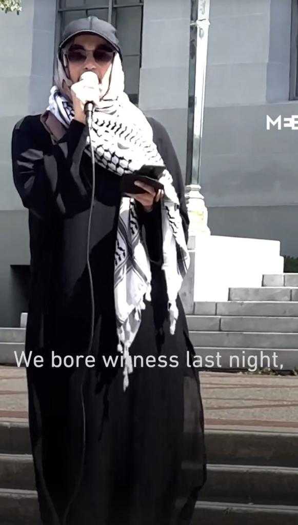Malak Afaneh in baseball cap and a hijab made out of a kefiyeh, and a long black robe, holding a microphone.