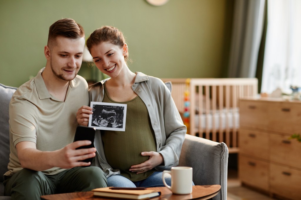 Young couple talking online on mobile phone and sharing their happiness with relatives showing them ultrasound image