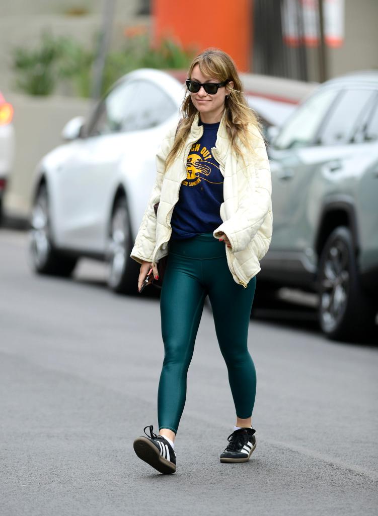 Olivia Wilde leaving her morning gym in Los Angeles, dressed in a cream jacket, graphic sweatshirt, green leggings, and wearing Adidas Samba sneakers